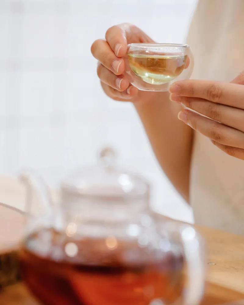 Close up of a person holding a small double walled glass cup of tea with a blurred glass teapot in the foreground.
