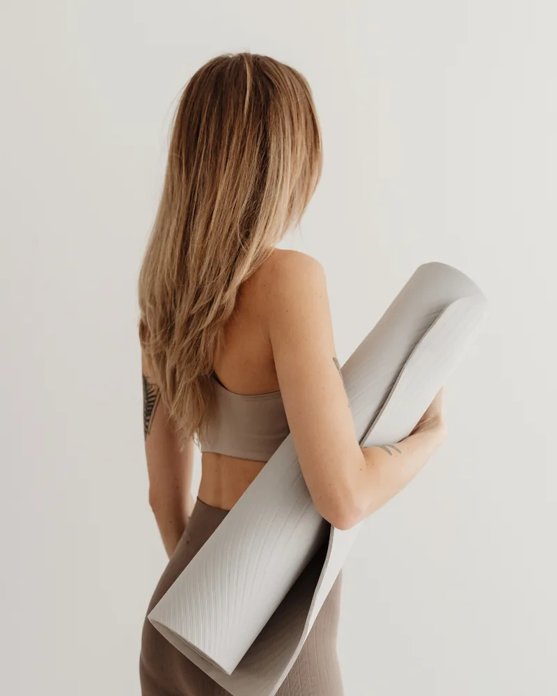 A woman viewed from behind, wearing a neutral-toned sports bra and leggings, holding a rolled-up light grey yoga mat under her arm against a plain white background.