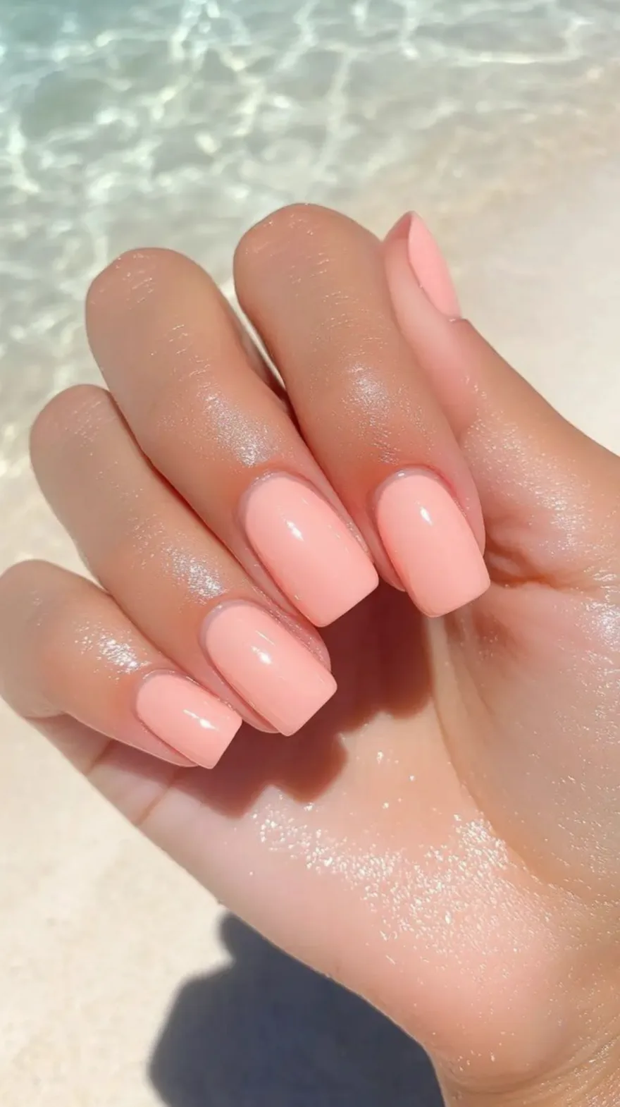 Close-up of a hand with glossy peachy-pink square nails held over sparkling tropical ocean water.