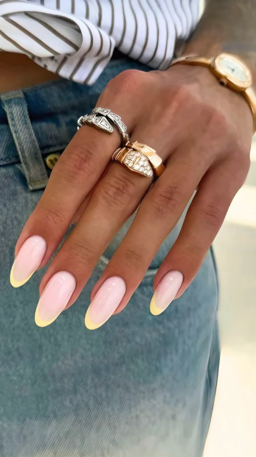 Almond shaped nails featuring a soft pink base with pale yellow French tips, worn with stacked gold and diamond rings.