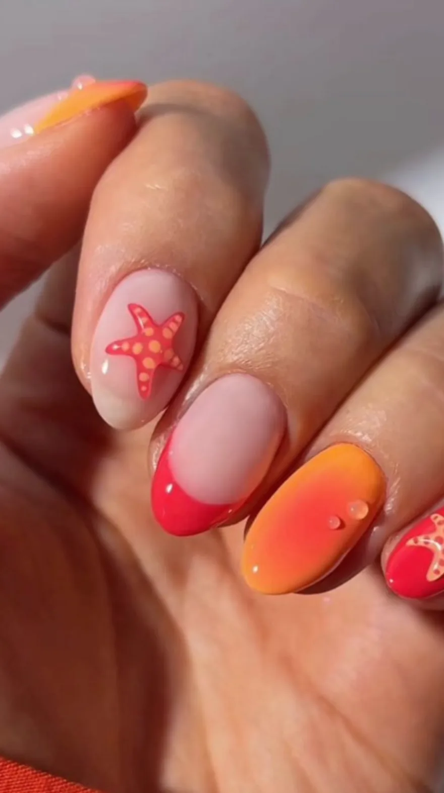 Close-up of almond nails featuring a mix of bright coral, orange ombre, and painted starfish details with small water droplet textures.