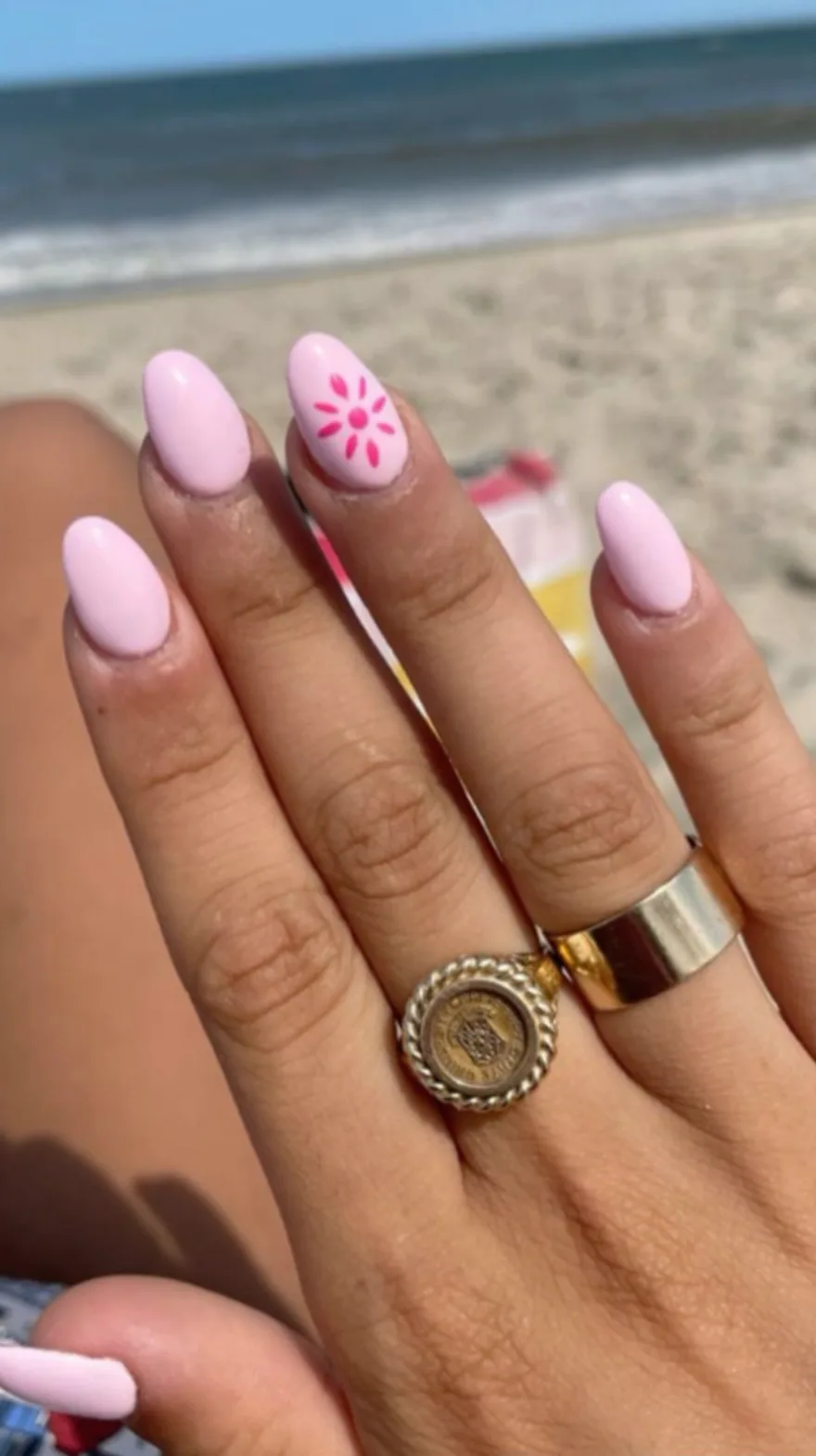 Soft pink almond nails against a sandy beach background, featuring one accent nail with a delicate painted pink flower.