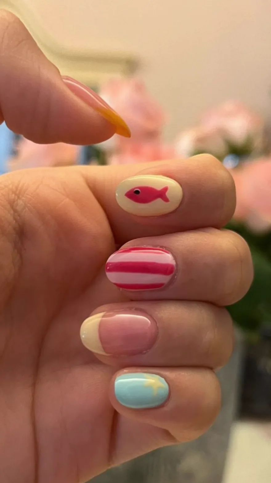 Hand holding a flower showing mix-and-match nail art including a pink fish, pink stripes, a yellow and pink french tip, and a blue base with a yellow starfish.
