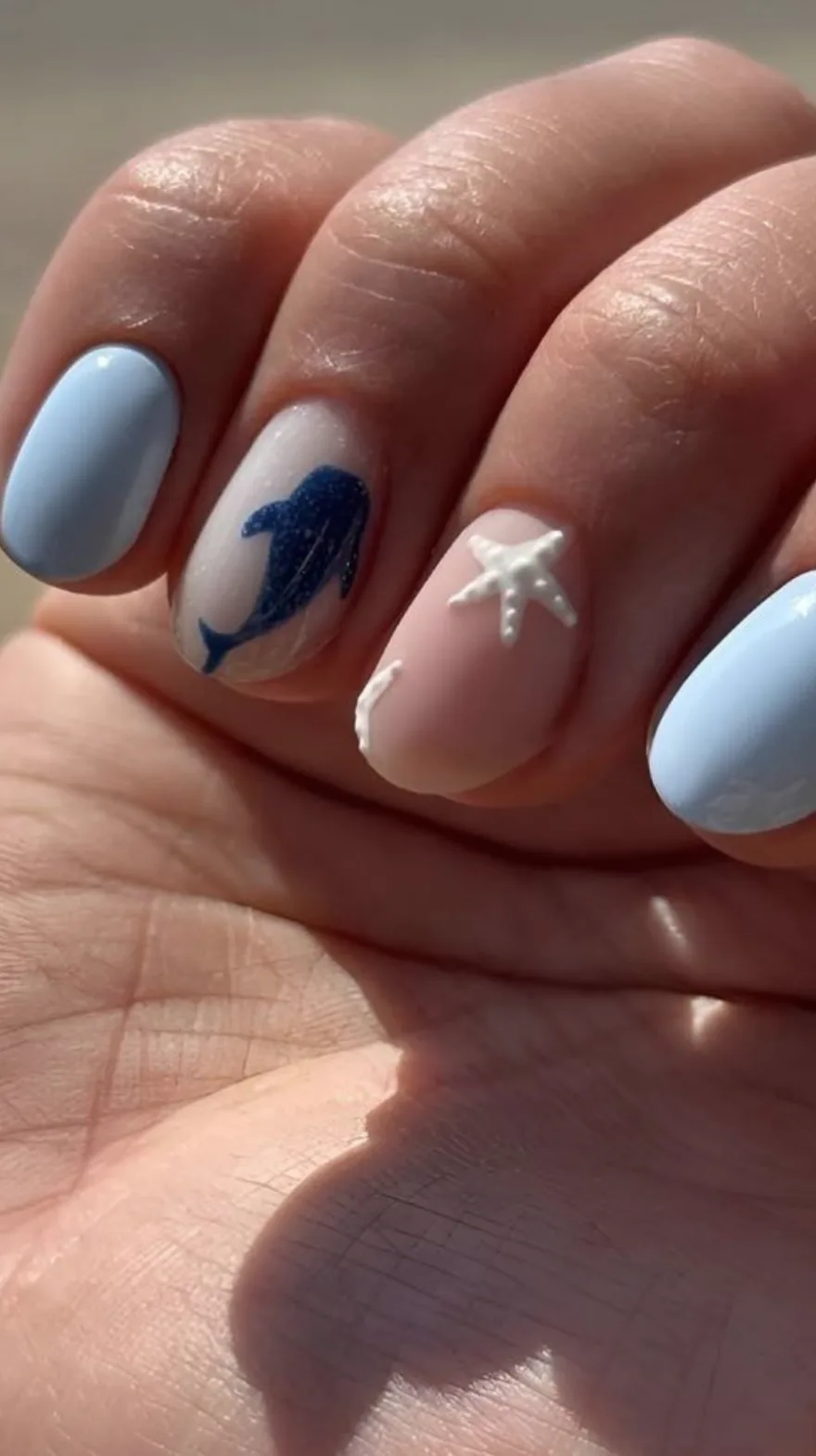 Hand resting on a leg showing almond nails featuring pale blue polish, white starfish and shell details, and a dark blue painted dolphin.