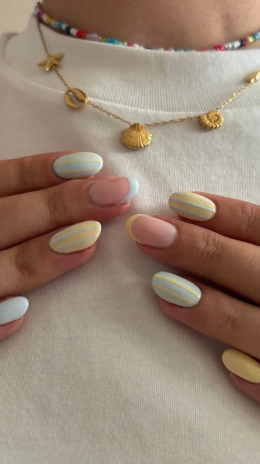 Oval nails painted with alternating soft pink bases, solid yellow, pale blue with yellow stripes, and yellow french tips, worn with a gold shell necklace.