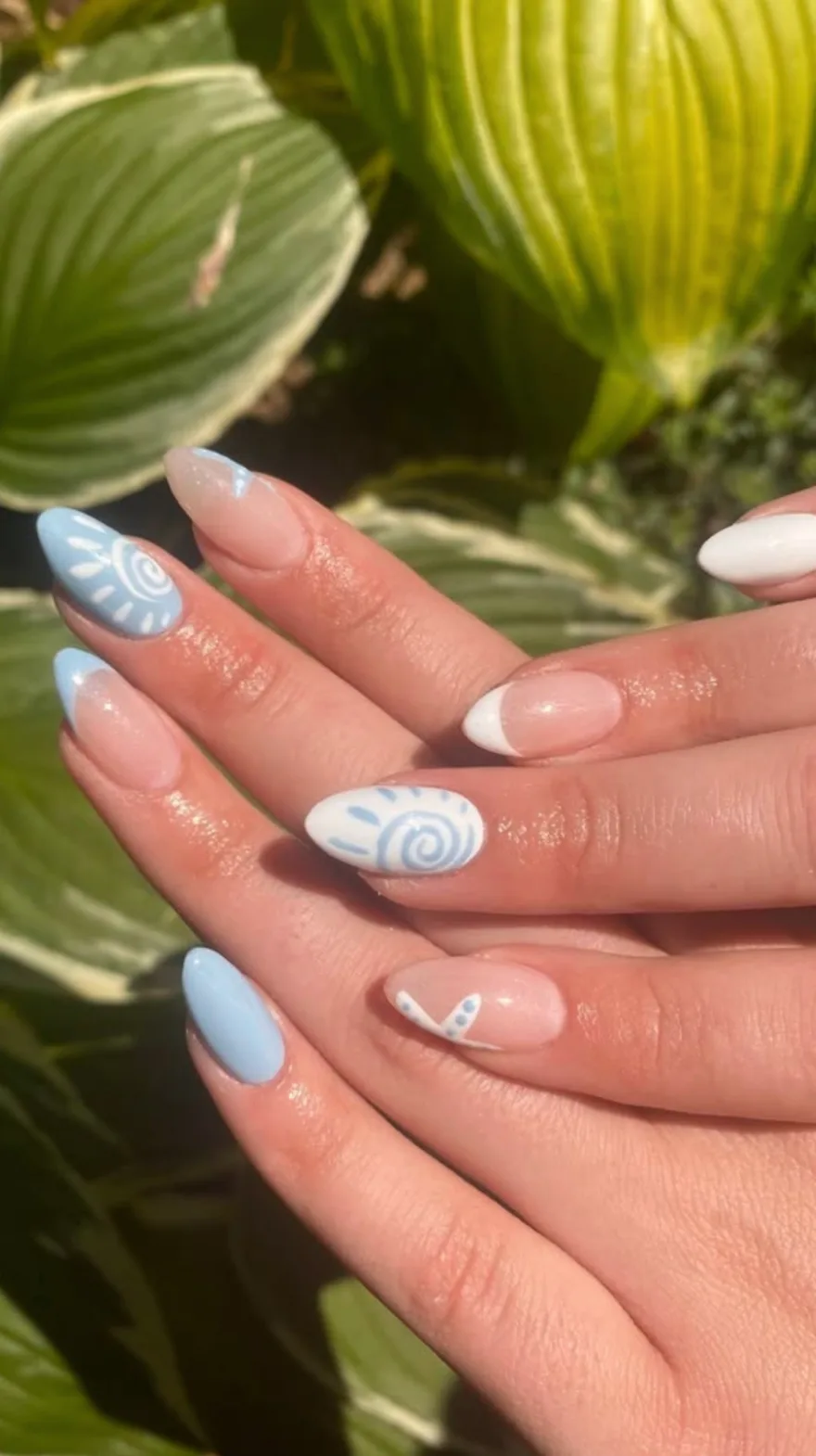 Hands resting against green leaves showing almond nails with pale blue polish, nude bases with white french tips, and delicate blue and white swirl patterns.