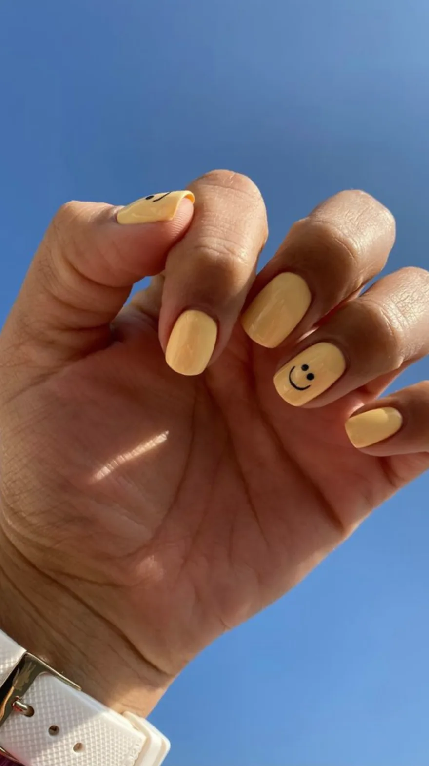 Hand with vibrant yellow short square nails held against a clear blue sky, featuring a small black smiley face on the ring finger.