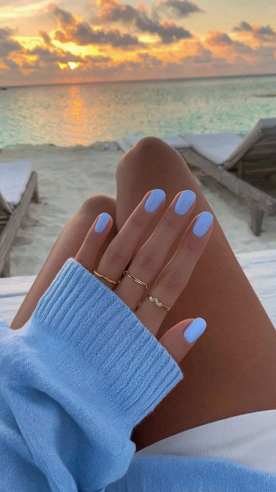 Creamy light blue nails on a hand resting against a blurred sunset beach background with a matching blue sweater.