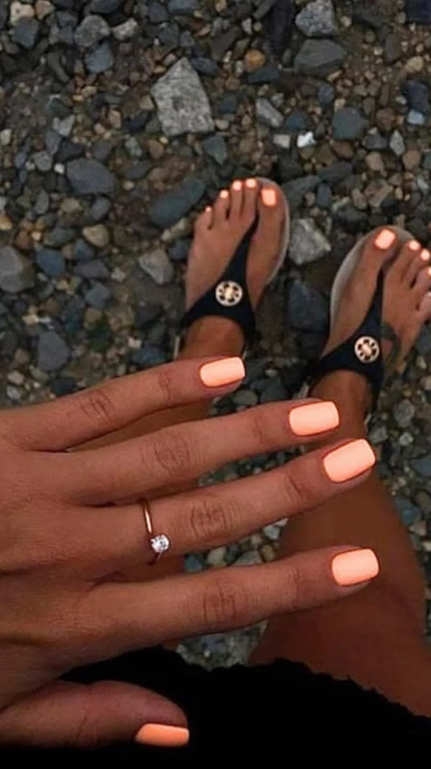 Hands and feet featuring matching bright peachy-coral polish on short square nails over a rocky beach background.