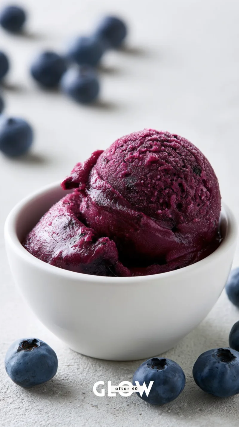 A close-up of a rich, velvety deep purple blueberry sorbet scoop resting inside a small white ceramic bowl, surrounded by fresh scattered blueberries.