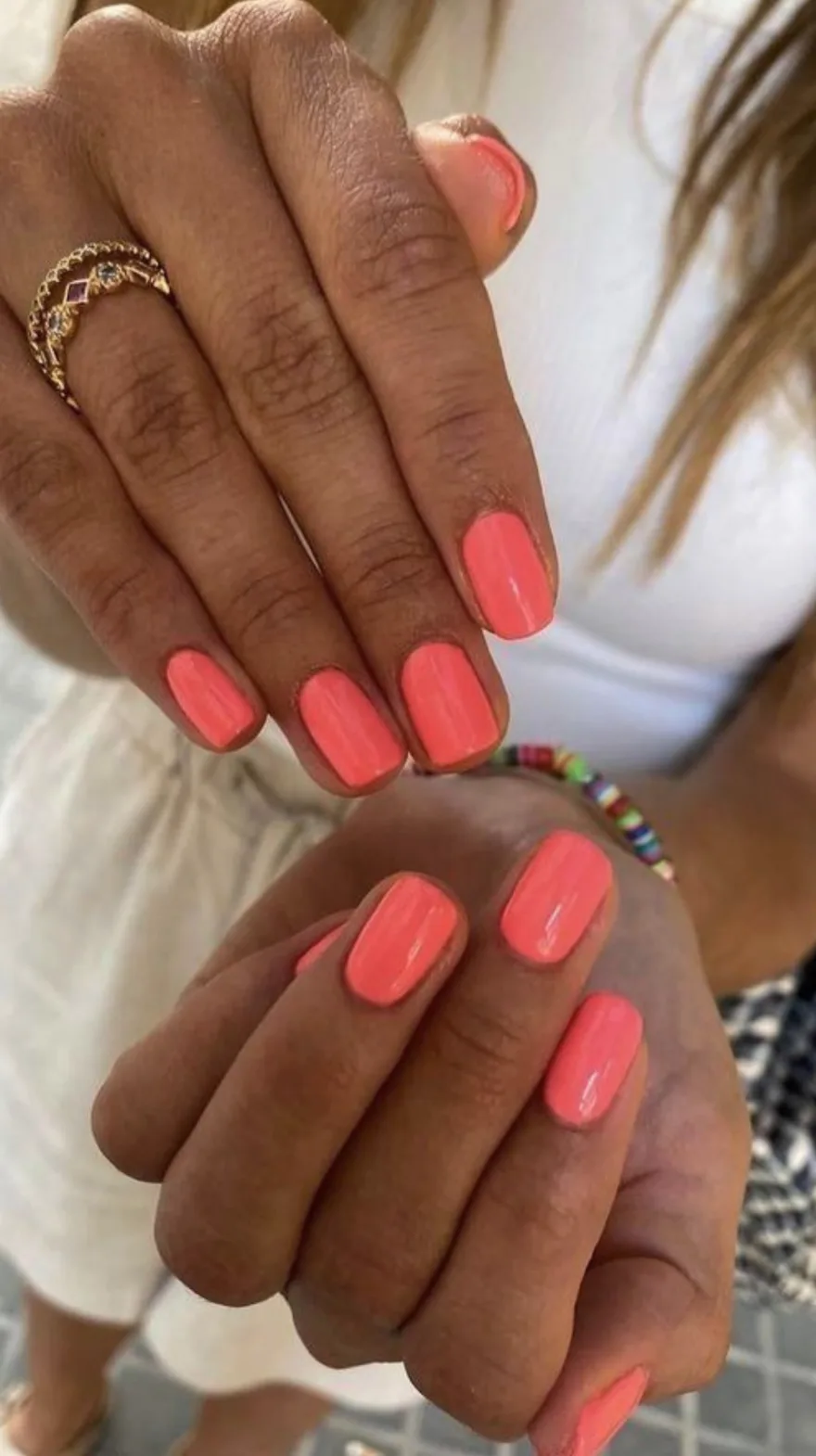 Vibrant coral-pink glossy nails on short square shapes held near a white linen outfit and colorful beaded bracelet.