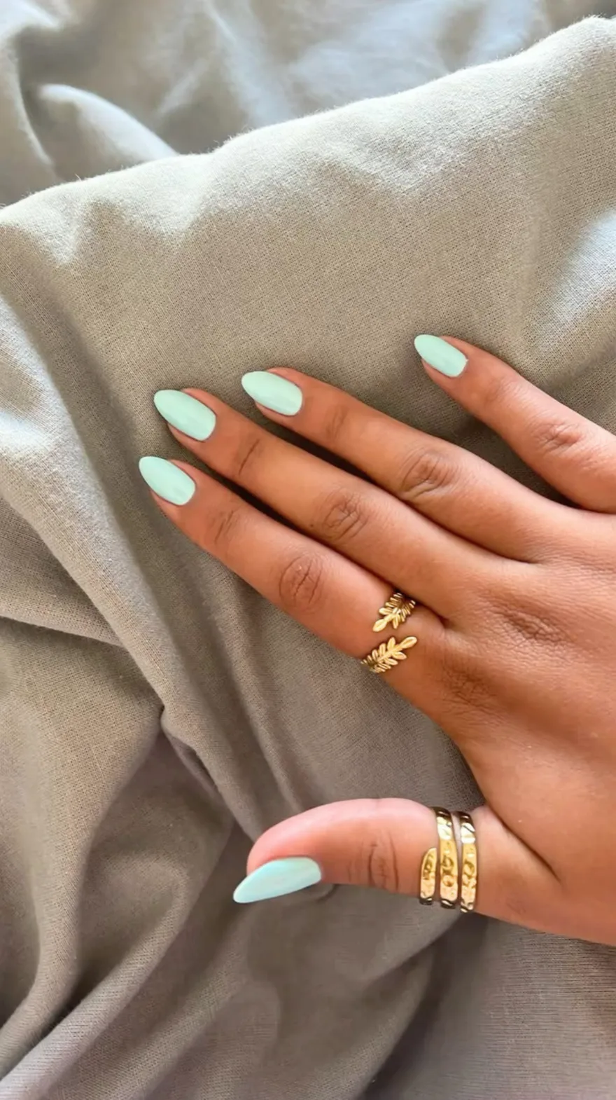 Pale mint green almond-shaped nails resting on a textured grey fabric with gold leaf-shaped rings.