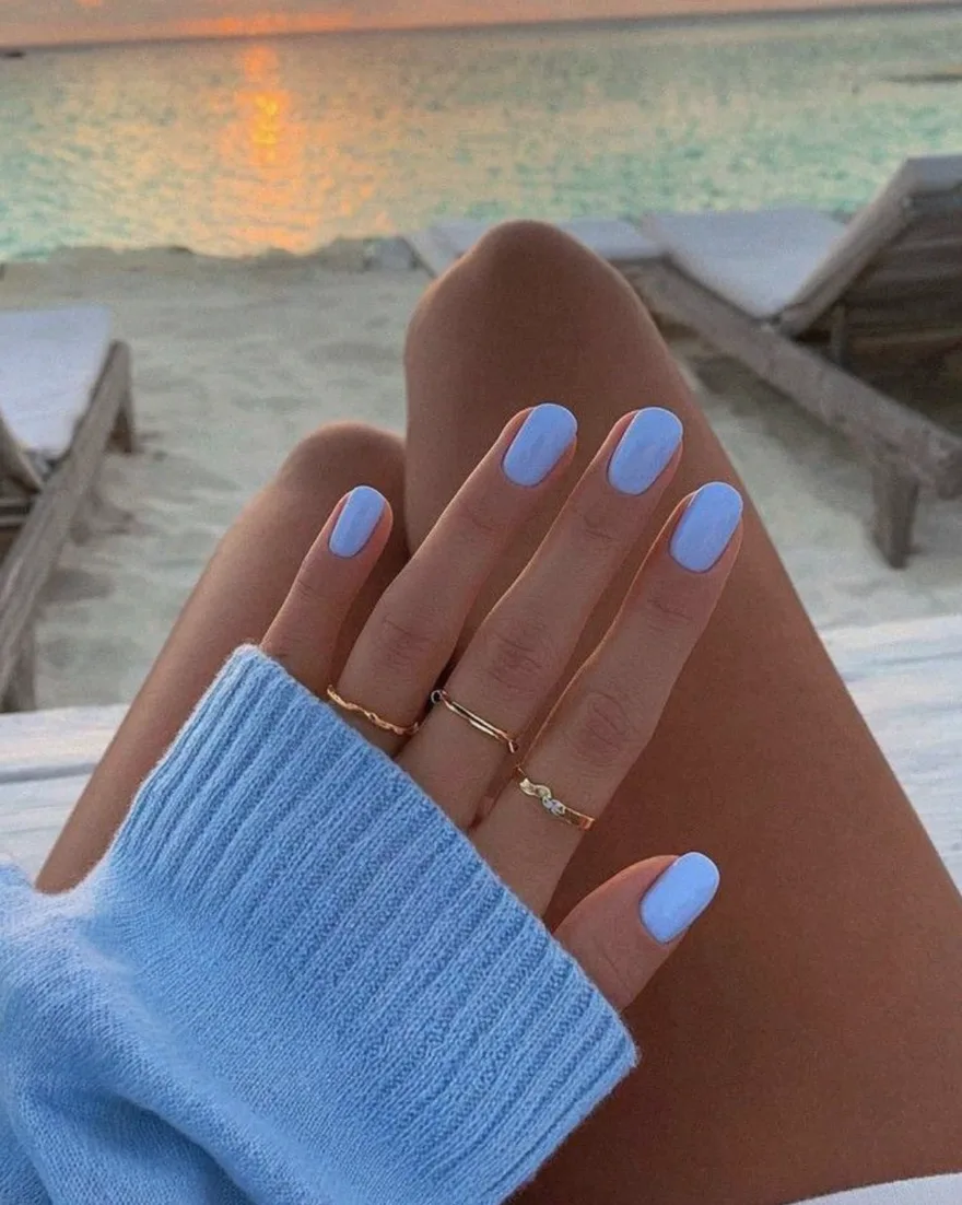 Hand with creamy sky blue almond nails resting against a blurred sunset beach background with gold rings and a light blue sweater sleeve.